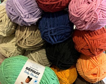 May include: A collection of colorful Bernat Baby Blanket yarn skeins in various shades, including green, purple, orange, and blue. The yarn is soft and plush, ideal for baby blankets and other craft projects. The label on the green skein reads "Bernat Baby Blanket".