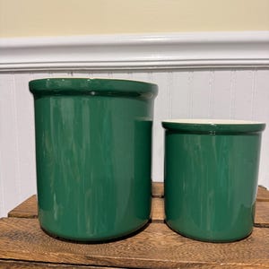 May include: Two green ceramic kitchen canisters with a glossy finish. The larger canister is taller than the smaller one. Both have a white interior and sit on a wooden surface.