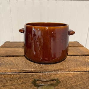 May include: A brown ceramic pot with two small handles. The pot has a glossy finish and sits on a wooden surface. The pot is a rich, warm brown color.