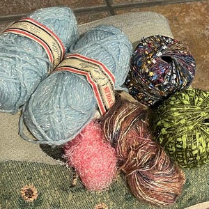 May include: Five balls of yarn in different colors and textures. Two balls are light blue with silver glitter, one is pink and fuzzy, one is brown and multi-colored, and one is green with gold glitter.