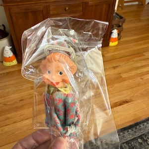 May include: A vintage doll with a red and green floral patterned outfit and a matching hat, packaged in a clear plastic bag. The doll has a rubber face with painted features and a long nose. The doll is a collectible item.