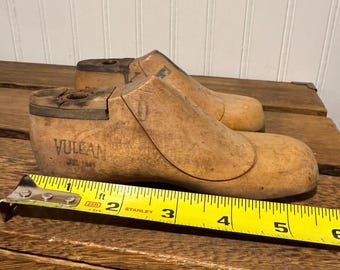 Op de afbeelding: Twee vintage houten schoenmallen, met het woord "VULCAN" en de datum "JUL. 1941" op één ervan gestempeld. Een gele rolmaat geeft aan dat de schoenmallen ongeveer 15 cm lang zijn. De schoenmallen liggen op een houten oppervlak.