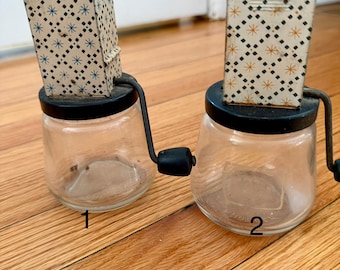 Op de afbeelding: Twee vintage glazen kruidenmolens met zwarte deksels en handvatten. Elke molen heeft een heldere glazen pot en een papieren bovenkant met patronen met blauwe en oranje sterrenpatronen. De molens zijn genummerd 1 en 2.