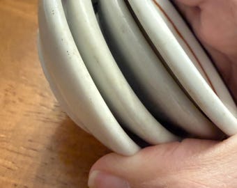 May include: A stack of four small, round, white ceramic dishes with curved edges. The dishes feature a smooth, glossy finish and a thin, dark-colored rim. The stack is held together, showing the side profile of the white ceramic material.