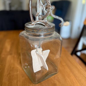 May include: An antique glass butter churn with a metal crank and gear mechanism. The churn has a clear glass jar with a wooden paddle inside. The metal components show signs of age and wear, giving it a vintage aesthetic.