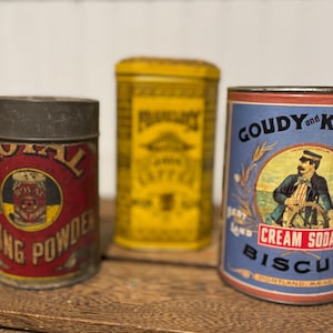 May include: Three vintage metal containers: a red and yellow Royal Baking Powder tin, a yellow tin labeled "Pharmacy Coffee", and a blue and white Goudy and Kent Cream Soda Biscuit tin. All are displayed on a wooden surface.