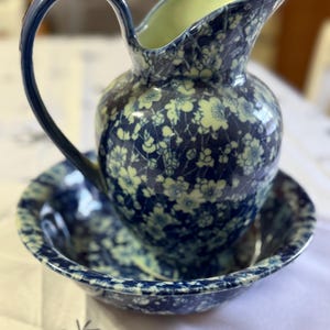 May include: A vintage blue and white floral pitcher and bowl set. The pitcher has a curved handle and a spout, and the bowl is round. The set is decorated with a delicate floral pattern in shades of blue and white, with a pale yellow interior.