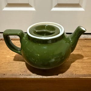 May include: A green ceramic teapot with a white rim and lid. The teapot has a curved handle and a spout. The lid has a small handle. The teapot is sitting on a wooden surface.