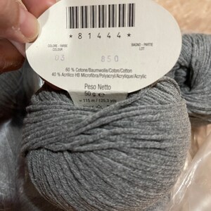 Op de afbeelding: Een bol grijs garen met een label waarop staat "Peso Netto 50 g e = 115 m / 125,3 yds". Het label bevat ook de tekst "60% Cotone/Baumwolle/Coton/Cotton 40% Acrilico HB Microfibra/Polyacryl/Acrylique/Acrylic".