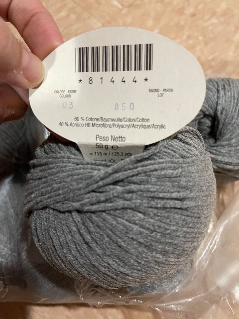 Op de afbeelding: Een bol grijs garen met een label waarop staat "Peso Netto 50 g e = 115 m / 125,3 yds". Het label bevat ook de tekst "60% Cotone/Baumwolle/Coton/Cotton 40% Acrilico HB Microfibra/Polyacryl/Acrylique/Acrylic".