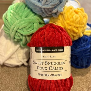May include: A collection of colorful yarn balls in shades of red, green, blue, and yellow. The front ball is red with a label that reads "Sweet Snuggles" and includes measurements of 109 yards and 8.8 ounces.