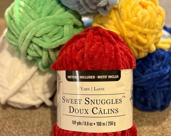 May include: A collection of colorful yarn balls in shades of red, green, blue, and yellow. The front ball is red with a label that reads "Sweet Snuggles" and includes measurements of 109 yards and 8.8 ounces.