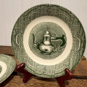 Puede incluir: Un plato vintage verde y blanco con una ilustración de tetera en el centro. El plato tiene un borde decorativo y se exhibe en un soporte rojo. El plato es de cerámica y forma parte de un juego de vajilla.