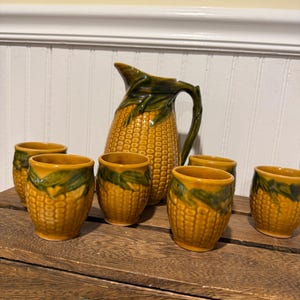 May include: A ceramic pitcher and six matching cups. The pitcher is shaped like an ear of corn, with a yellow body and green leaves. The cups are also corn-shaped, with a yellow body and green accents. The set is displayed on a wooden surface.
