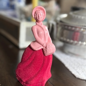 May include: A vintage figurine of a woman in a long red dress and a pink hat and jacket. The figurine has a textured, velvet-like appearance. The woman is holding a small muff. The background is blurred, with a wooden table in the foreground.