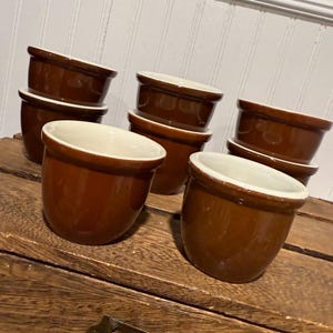 May include: A collection of brown ceramic ramekins with white interiors. The ramekins are arranged on a wooden surface, showcasing their rounded shape and contrasting colors. The image highlights the simple, classic design of the bakeware.