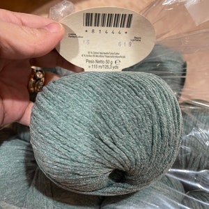 May include: A close-up of a ball of teal yarn with a label. The label includes the text "* 8 1 4 4 4 *", "15", "616", and "Peso Netto 50 g = 115 m/125.3 yds". The yarn is made of 50% cotton and 40% acrylic.