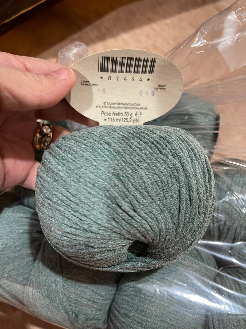 May include: A close-up of a ball of teal yarn with a label. The label includes the text "* 8 1 4 4 4 *", "15", "616", and "Peso Netto 50 g = 115 m/125.3 yds". The yarn is made of 50% cotton and 40% acrylic.