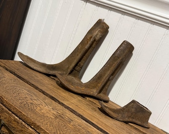 Op de afbeelding: Drie antieke, roestige metalen schoenmallen van verschillende maten liggen op een houten oppervlak. De mallen zijn bruin en vertonen tekenen van ouderdom en slijtage. De achtergrond is een witte wand met panelen.