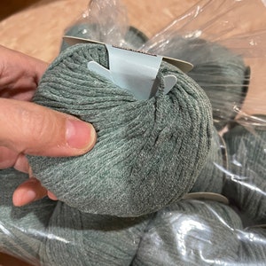 May include: A close-up of several balls of sage green yarn, partially visible through a clear plastic bag. The yarn is tightly wound, showing a textured surface. The image suggests a craft or knitting project.