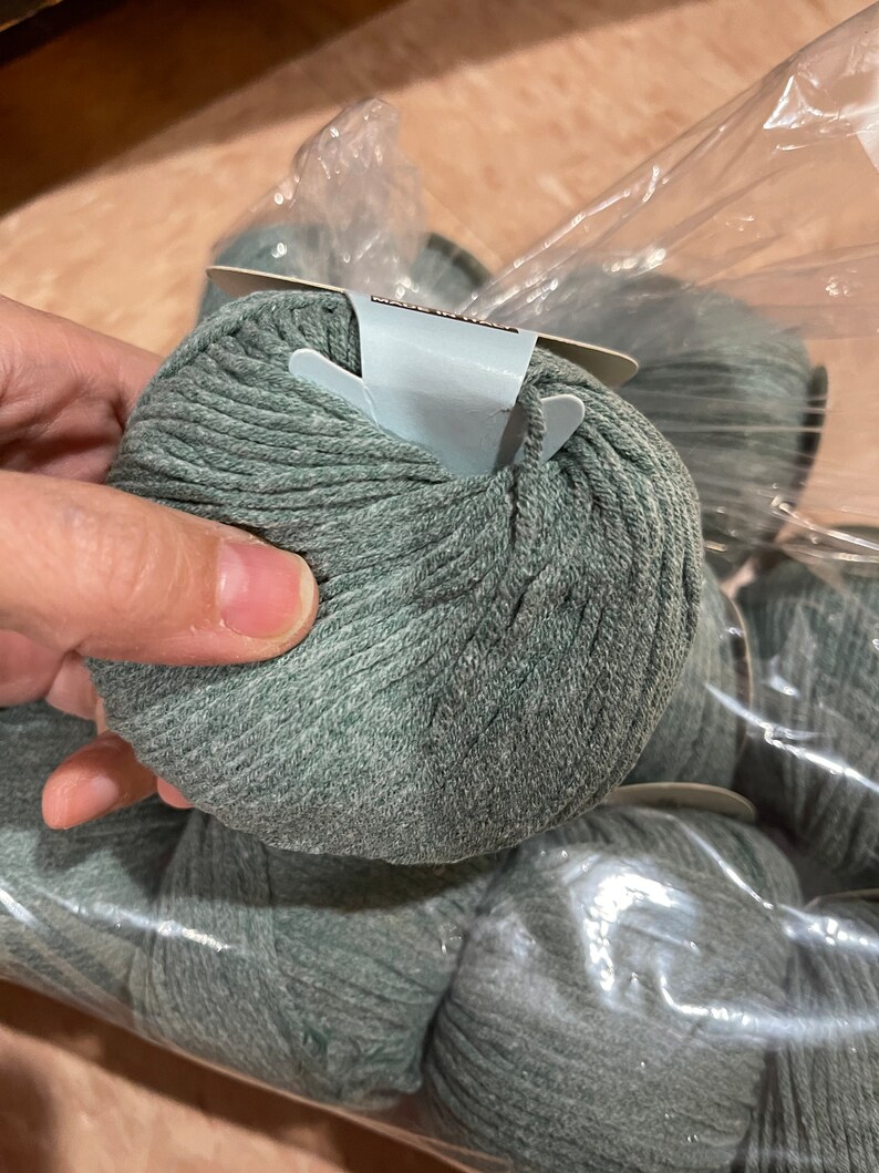 May include: A close-up of several balls of sage green yarn, partially visible through a clear plastic bag. The yarn is tightly wound, showing a textured surface. The image suggests a craft or knitting project.