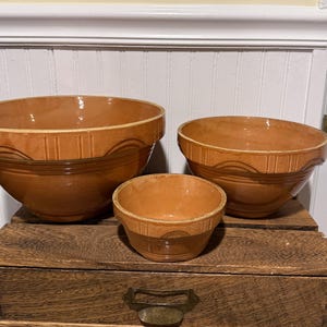 Peut inclure: Trois saladiers en céramique marron de différentes tailles. Les bols présentent un motif festonné sur le bord et une bande décorative près de la base. Ils sont disposés sur une surface en bois.