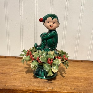May include: A ceramic figurine of a seated elf wearing a green suit and a green cap with a red pom-pom. The elf is positioned atop a green base surrounded by a decorative ring of artificial holly leaves and small red berries.