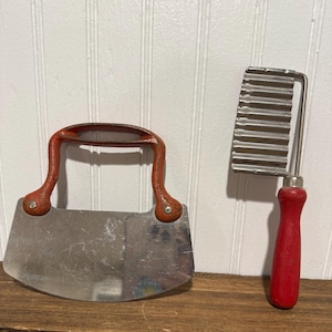 May include: Two vintage kitchen tools: a curved, stainless steel mezzaluna knife with a red handle and a crinkle cutter with a red handle. Both tools are on a wooden surface against a white paneled wall.
