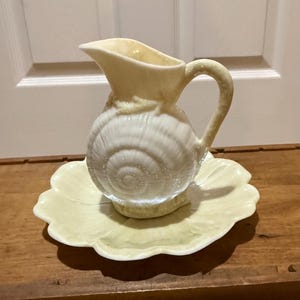 Könnte beinhalten: Eine cremefarbene Keramikkrug mit einem Muscheldesign, die auf einer passenden Platte mit gewelltem Rand steht. Der Krug hat einen gebogenen Griff und einen Ausguss. Das Set ist in einem weichen Gelb gehalten und ist wahrscheinlich ein Dekorationsartikel.