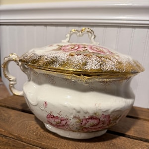 Op de afbeelding: Antieke witte porseleinen serveerschaal met deksel, gouden rand en roze bloemmotieven. De schaal heeft een decoratieve handgreep en een geschulpte rand. Een vintage stuk.