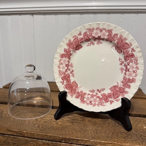 May include: A white plate with a pink floral design and a ruffled edge, displayed on a black stand. A clear glass dome is to the left. The plate features a pattern of pink flowers and leaves.