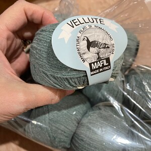 May include: A package of gray-green yarn balls. The label reads "VELLUTE FILATI DI ROMAGNO SESIA MANIFATTURA" with a drawing of a bird. The label also says "MAFIL MADE IN ITALY".