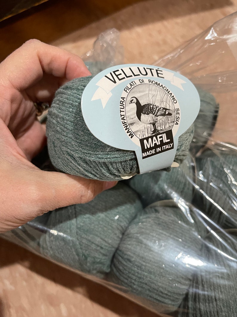 May include: A package of gray-green yarn balls. The label reads "VELLUTE FILATI DI ROMAGNO SESIA MANIFATTURA" with a drawing of a bird. The label also says "MAFIL MADE IN ITALY".