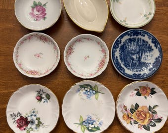 May include: A collection of nine small ceramic dishes arranged in a grid. The set includes various round and oval plates featuring floral patterns, gold-trimmed edges, and one blue and white plate with a deer graphic and the text Wisconsin.