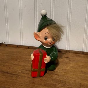 May include: A ceramic elf figurine wearing a dark green outfit and a matching pointed hat with a white pom-pom. The elf has light brown hair and is holding a rectangular red gift box decorated with a gold ribbon and a gold cross pattern.