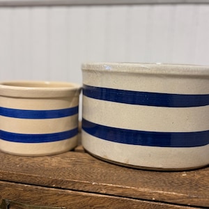 May include: Two antique stoneware crocks with blue horizontal stripes. The crocks are cylindrical, one smaller than the other. The smaller crock is on the left, and the larger crock is on the right. The crocks are beige with dark blue stripes.