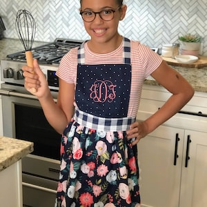May include: A young person wearing a blue and white gingham and floral print apron with a pink monogram. The apron has a white scalloped edge. The person is holding a whisk in their right hand.