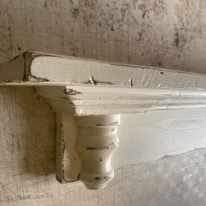 May include: A distressed, off-white wooden shelf with a decorative corbel support. The shelf has a slightly weathered appearance with visible wood grain and dark accents, suggesting a vintage or farmhouse style. The shelf is likely for home decor.