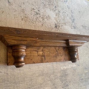 May include: A rustic, wooden shelf with a warm brown finish. The shelf features a rectangular design with two decorative, turned supports. The wood grain is visible, adding to the shelf's natural aesthetic. Perfect for displaying items.