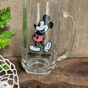May include: Clear glass Mickey Mouse mug with a handle. The mug features a full-body illustration of Mickey Mouse in his classic outfit, with the words "Mickey Mouse" printed below. The mug is a rectangular shape.