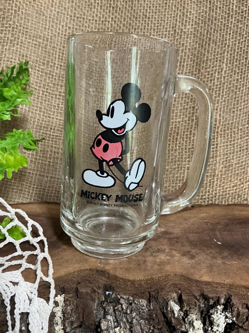 May include: Clear glass Mickey Mouse mug with a handle. The mug features a full-body illustration of Mickey Mouse in his classic outfit, with the words "Mickey Mouse" printed below. The mug is a rectangular shape.