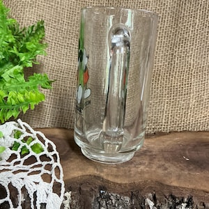 May include: Clear glass mug with a handle. The mug features a colorful graphic of a cartoon character. The mug is sitting on a wooden surface. The background is a burlap material.