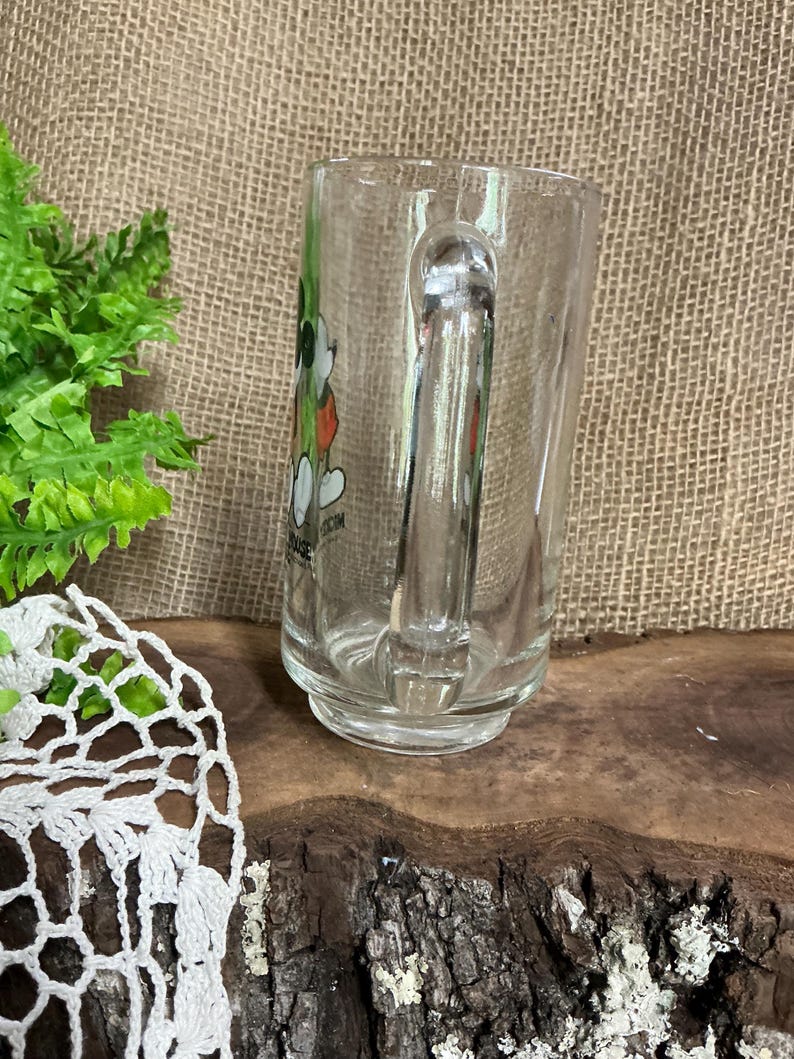 May include: Clear glass mug with a handle. The mug features a colorful graphic of a cartoon character. The mug is sitting on a wooden surface. The background is a burlap material.