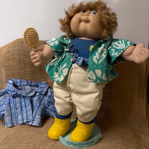 Puede incluir: Una muñeca Cabbage Patch Kids con una camisa azul, pantalones color crema y una camisa con estampado floral verde y blanco. La muñeca tiene botas amarillas y un peine. También hay una segunda camisa a cuadros azul y blanco.