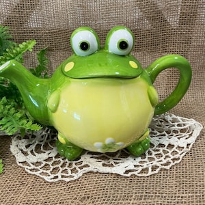 May include: A ceramic teapot shaped like a frog with a bright green body, a pale yellow belly, and two large protruding eyes on the lid. The teapot features a curved handle, a spout on the left side, and a small white flower detail on the lower front.