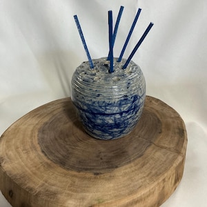 Op de afbeelding: Een keramische vaas met een blauw en wit gemarmerd ontwerp, met verschillende blauwe rietjes. De vaas staat op een ronde, natuurlijke houten schijf. De vaas is ongeveer 10 cm hoog en 8 cm breed.