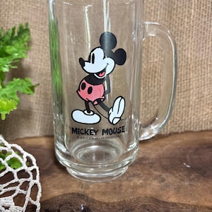 May include: Clear glass mug featuring Mickey Mouse. The cartoon character is depicted in black, white, and red, with the text "Mickey Mouse" and "Walt Disney Productions" below. The mug has a handle and a slightly flared base.
