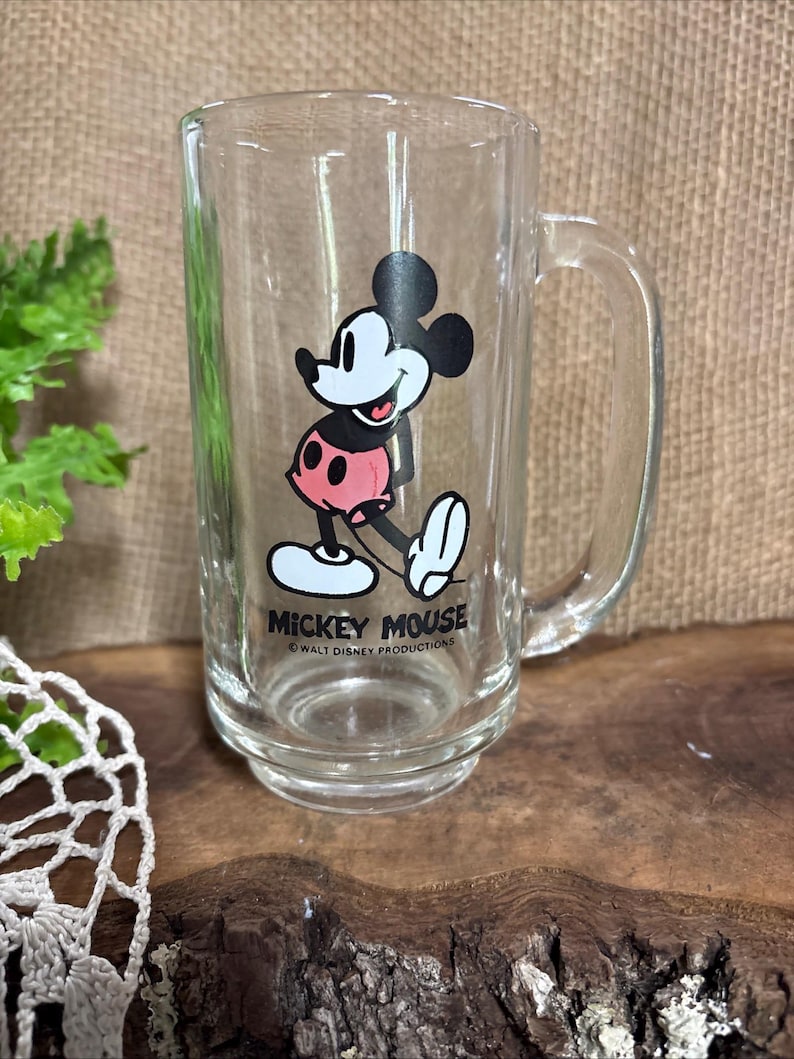 May include: Clear glass mug featuring Mickey Mouse. The cartoon character is depicted in black, white, and red, with the text "Mickey Mouse" and "Walt Disney Productions" below. The mug has a handle and a slightly flared base.
