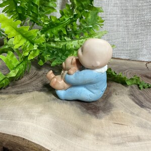 Pode incluir: Uma figura de cerâmica de um bebê em um macacão azul claro, segurando um pequeno ursinho de pelúcia. O bebê tem uma tez clara e está sentado. A figura está em uma superfície de madeira com folhagem verde ao fundo.