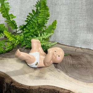 May include: A ceramic baby figurine, lying on its back with arms raised. The baby is wearing a white diaper with a gold pin. The figurine has a light skin tone and rosy cheeks. The baby is resting on a wooden surface with a green fern in the background.